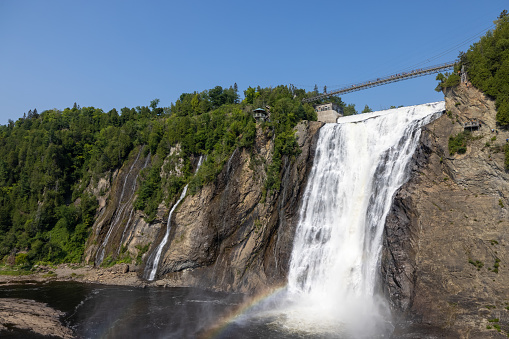 Montmorency Falls in Summer, Quebec City, Canada. It is a beautiful sunny summer day and there is a rainbow. Tourists are walking on the suspension bridge.