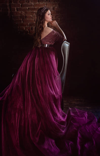 piękna młoda ładna urocza księżniczka w długiej fioletowej sukience królowej i koronie, z długimi włosami i makijażem w minimalistycznym wnętrzu na poddaszu w pobliżu skórzanego krzesła - women crown princess 20s zdjęcia i obrazy z banku zdjęć