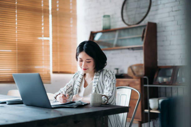 istock Concentrated young Asian woman working from home using laptop computer and handling paperworks in the living room at home. Flexible lifestyle. Remote working concept 1367469119