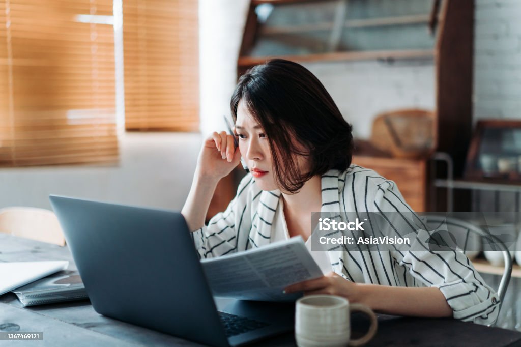 Young Asian woman handling home finances with laptop, looking worried while going through financial bills. Financial plan, tax, spending and budgets, financial problems concept People Stock Photo
