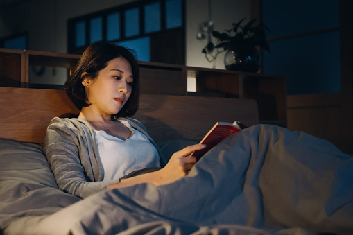 Relaxed young Asian woman lying in bed and reading a book at night