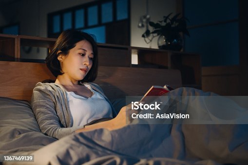 istock Relaxed young Asian woman lying in bed and reading a book at night 1367469139