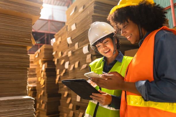 ingeniera asiática y africana con casco de seguridad y chaleco sosteniendo portapapeles y tome nota en el papel en el almacén de piezas automotrices. productos y cartón ondulado. fábrica para la fabricación y procesamiento de papel. - industry asian ethnicity asia manager fotografías e imágenes de stock