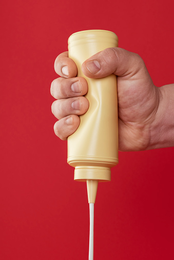 Man hand squeezing a bottle of mayo against a red-colored background. Pouring mayonnaise from a plastic bottle.