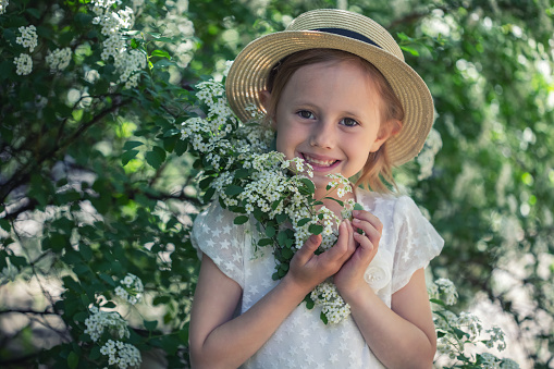 Portrait of beautiful caucasian little girl in boho dress and a hat in blooming bush with white flowers of spirea with selective focus and soft focus.