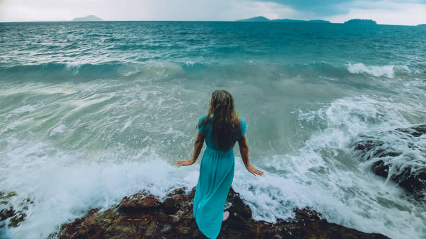 Woman meditates, relaxes on a rock reef hill in stormy morning rain cloudy sea. Concept feminine, relax, sexual health. Dark dramatic silhouette view stock photo