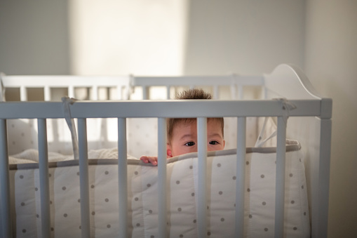 Cute six months old baby boy peeking through his crib fence in the early morning to wake up his parents and play in the bedroom