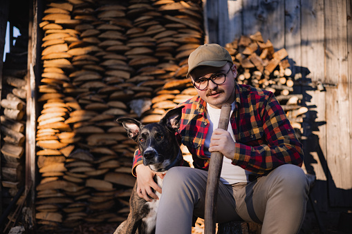 Portrait of a young Caucasian male lumberjack sitting in front of his wooden house in the forest, holding an axe and taking a break from chopping up the wood while hugging his pet dog