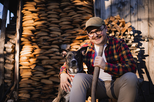 Portrait of a young Caucasian male lumberjack sitting in front of his wooden house in the forest, holding an axe and taking a break from chopping up the wood while hugging his pet dog and smiling