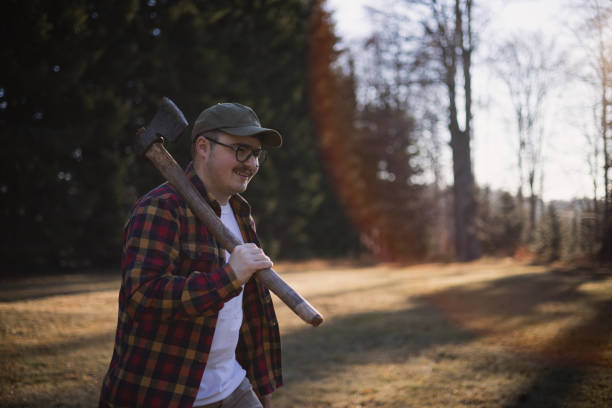 Young Caucasian male lumberjack walking into the forest to gather wood for the fire, carrying the axe and smiling