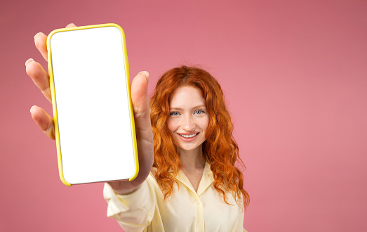 Excited redhead girl holding big smartphone with white blank screen in hand, showing close to camera. Gadget with empty free space for mock up, banner isolated on pink background. High quality photo