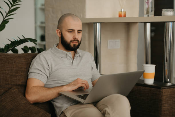istock A bald man with a beard works remotely on his laptop sitting on a sofa at home. 1367514037