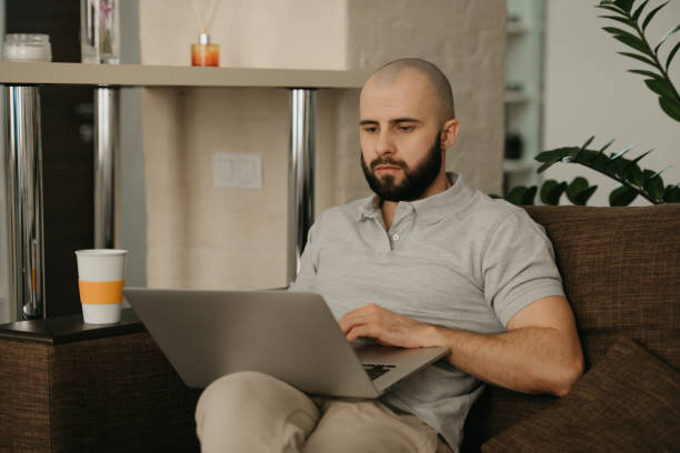 istock A bald man with a beard works remotely on his laptop sitting on a sofa at home. 1367514038