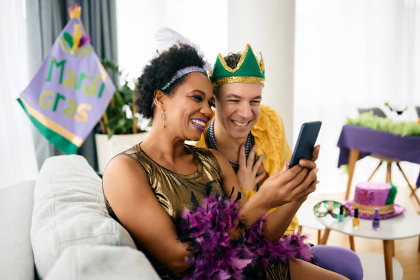 Carefree multi-ethnic friends using smart phone during Mardi Gras party at home. Happy African American woman and her male friend in Mardi Gras costumes using mobile phone at home party during the festival. carnival mask women party stock pictures, royalty-free photos & images