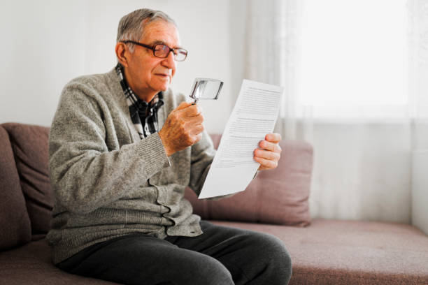 senior man holding an magnifying glass he uses to help him read because he suffers from wet macular degeneration. - macular degeneration imagens e fotografias de stock