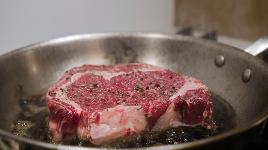 One juicy rib eye beef steak frying in high fat, in a pan. Red meat spiced with pepper, close up shot.