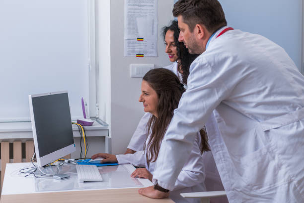 istock Two nurses ( female physicians ) and a male doctor standing next to a pc, mac ( desktop computer ) in the doctor's office while discussing a medical report. 1367542622
