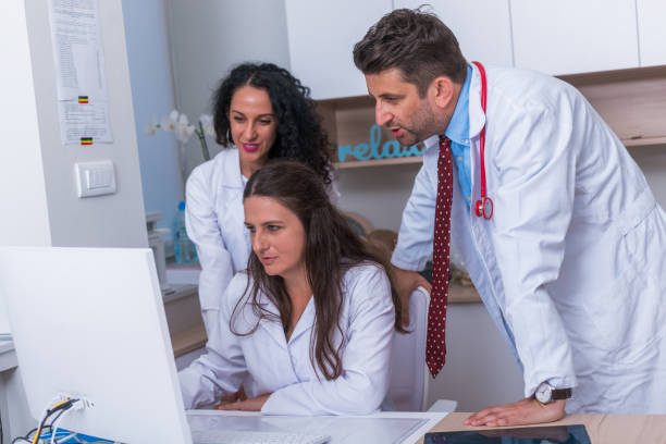 istock Two nurses ( female physicians ) and a male doctor standing next to a pc, mac ( desktop computer ) in the doctor's office while discussing a medical report. 1367542626