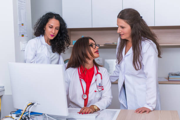 istock ( Medical Team ) Female doctor ( medic ) in her 30's working on her computer while two female nurses stand next to her and discuss a patients illness. 1367542634