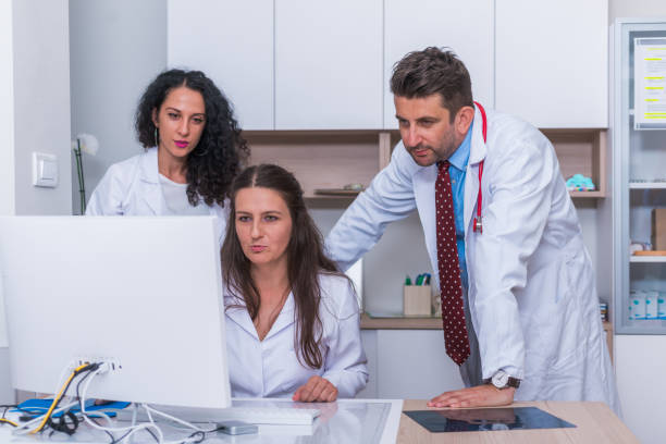 istock Two nurses ( female physicians ) and a male doctor standing next to a pc, mac ( desktop computer ) in the doctor's office while discussing a medical report. 1367542644