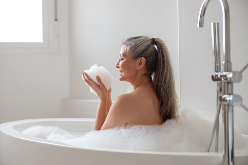 Smiling woman in beautiful bright bathroom holding white foam in hands while sitting and taking a bath