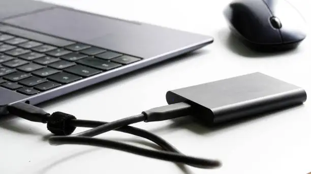 An external portable SSD drive connected to a laptop on a white background next to a computer mouse. Portable Solid State Drive. The concept of modern gadgets and the use of an SSD disk. Close-up