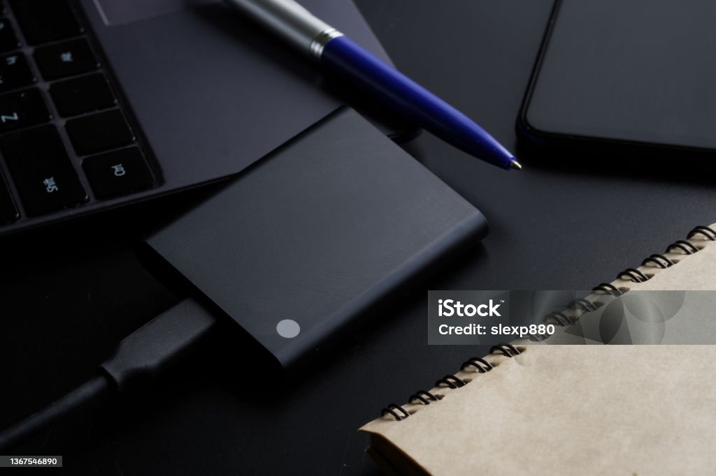 External portable SSD disk on a black background next to a laptop, smartphone, notepad and pen. Solid State Drive. The concept of modern technologies, gadgets and the use of an SSD disk External portable SSD disk on a black background next to a laptop, smartphone, notepad and pen. Solid State Drive. The concept of modern technologies, gadgets and the use of an SSD disk. Close-up Spatholobus Suberectus Dunn Stock Photo