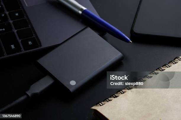 External Portable Ssd Disk On A Black Background Next To A Laptop Smartphone Notepad And Pen Solid State Drive The Concept Of Modern Technologies Gadgets And The Use Of An Ssd Disk Stock Photo - Download Image Now