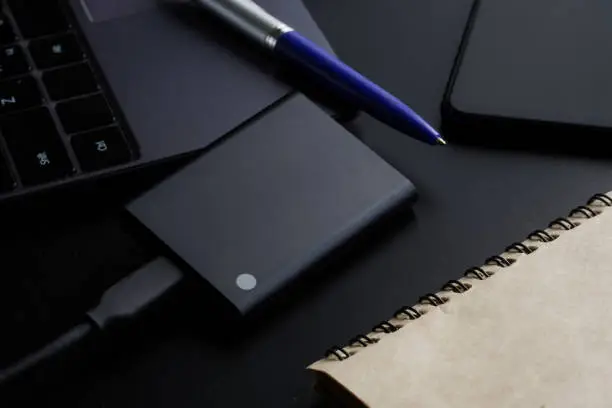 External portable SSD disk on a black background next to a laptop, smartphone, notepad and pen. Solid State Drive. The concept of modern technologies, gadgets and the use of an SSD disk. Close-up