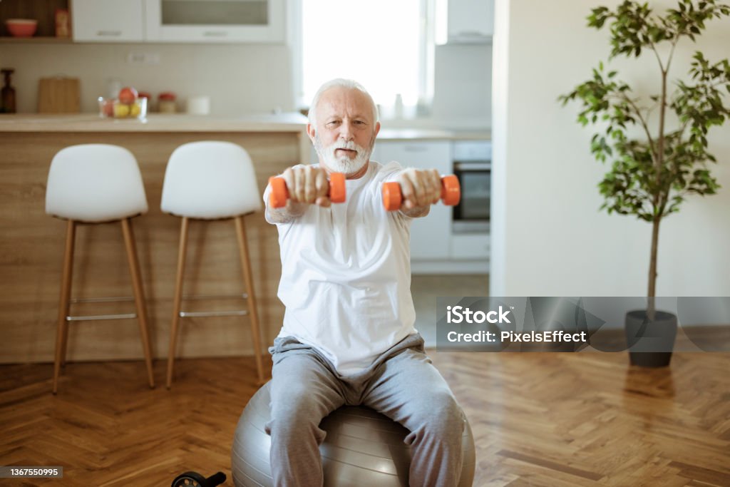 Senior Man Doing Exercises With Dumbbells at Home Bearded senior man doing exercises with dumbbells sitting on fitball stretching at home indoor Domestic Life Stock Photo