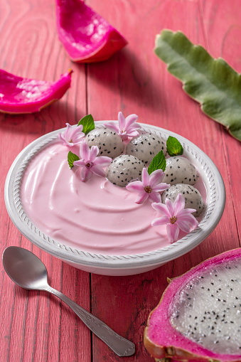 Pitahaya and strawberry yogurt breakfast bowl with mint leaves and pink flowers decoration on pink wood table, dragonfruit pitaya. Flowers are shown as a decorative suggestion, not such an edible part of the recipe.