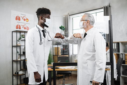 Side view of senior caucasian doctor greeting his african american colleague with elbow bumping during coronavirus pandemic. Two medical worker wearing face masks and lab coats.
