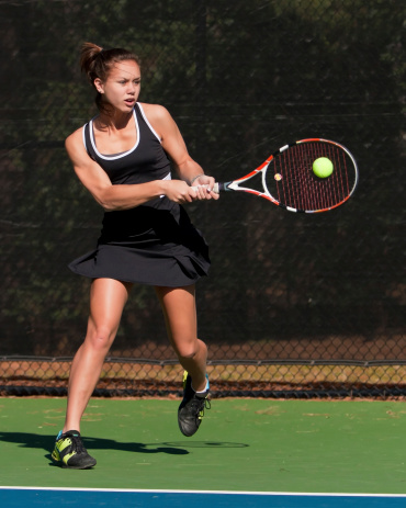 Very fit girl teenage tennis player smacks backhand, ball is frozen on racket head.