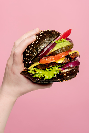 Veggie burger made with a blueberry infused, egg free brioche bread roll, filled with hummus, salad leaves, red onions, bean and lentil burger, vegan cheese, red pepper, avocado, pickles and tomatoes. Photographed against a pink background with a teenage girl’s hand holding the burger. Colour, vertical format with some copy space.