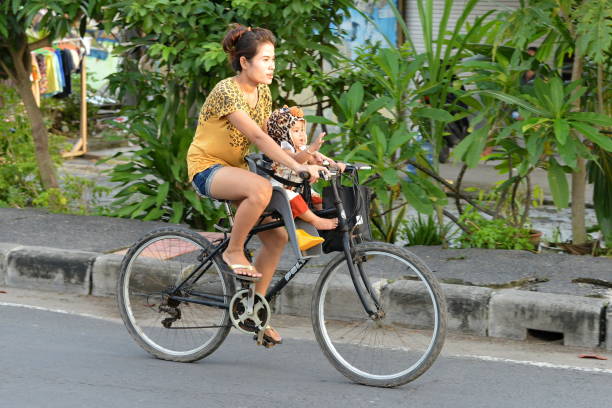 Bali, Indonesia - March 16, 2014: A woman rides a bicycle with a child passenger on a city street.