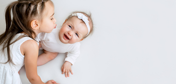 Six-month-old baby sobs avoiding the kisses of his little sister. Relationship of children in a family with an age difference. Girls in white clothes on a white studio background with a copy space