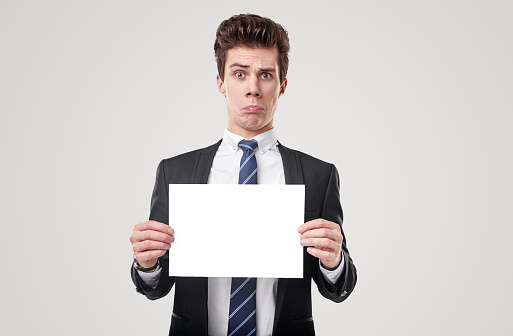 Young funny man in formal suit and tie holding empty white poster and looking with amazement at camera on white background