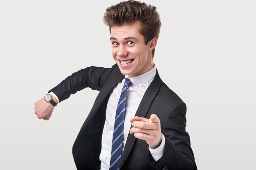 Comic punctual male entrepreneur in suit pointing at camera with forefinger and showing wristwatch for concept of time management on gray background in studio