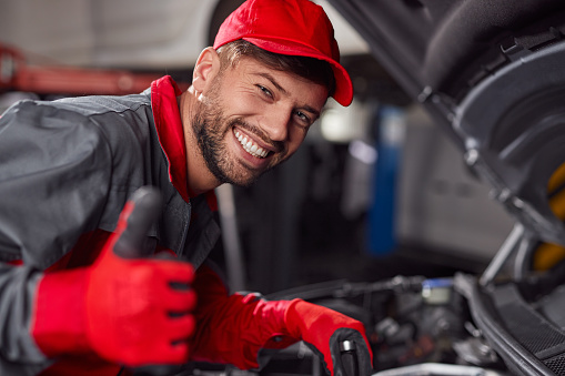 Cheerful male technician in uniform fixing broken automobile and showing like gesture while looking at camera and working in car service