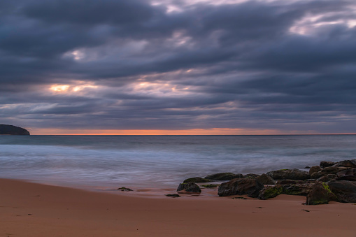 Sunrise seascape with rocks on the beach and low heavy clouds from Killcare Beach on the Central Coast, NSW, Australia.