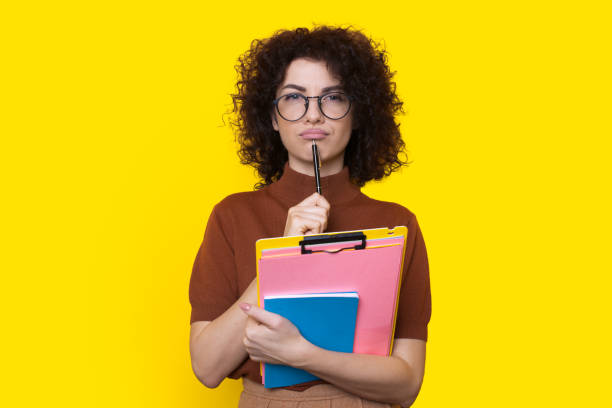 istock Curly woman with a note in hand thinking about what homework to do for tomorrow isolated over yellow background. Curly haired student. 1367600267