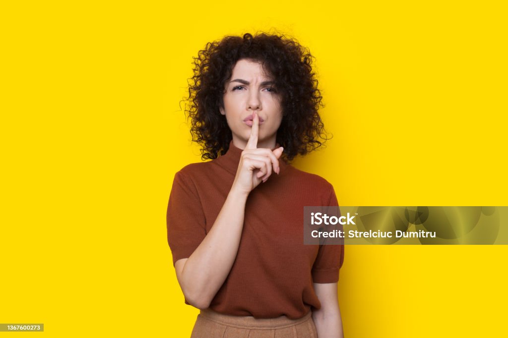 Portrait of serious woman showing silence gesture with finger over lips isolated over yellow background. Shh sign. Curly hair. Curly Hair Stock Photo