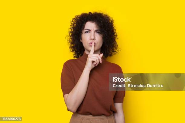 Portrait Of Serious Woman Showing Silence Gesture With Finger Over Lips Isolated Over Yellow Background Shh Sign Curly Hair Stock Photo - Download Image Now