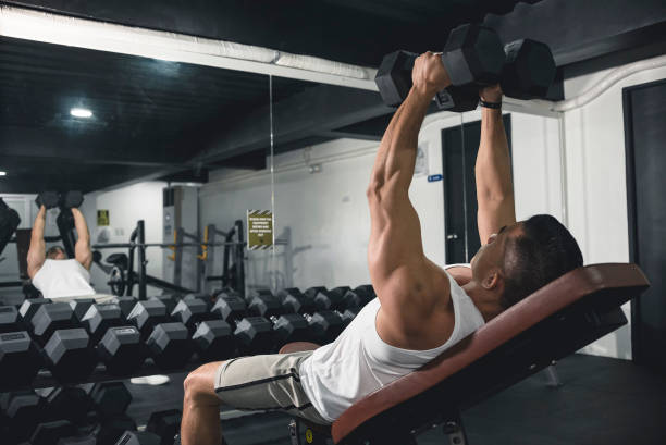 un uomo in canottiera bianca fa alcune mosche manubrio inclinate. allenarsi, allenare il torace con un esercizio di isolamento. - weightlifting foto e immagini stock