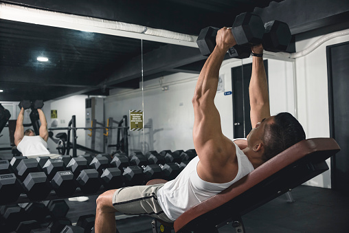 A man in white tank top does some incline dumbbell flys. Working out, training chest with an isolation exercise.