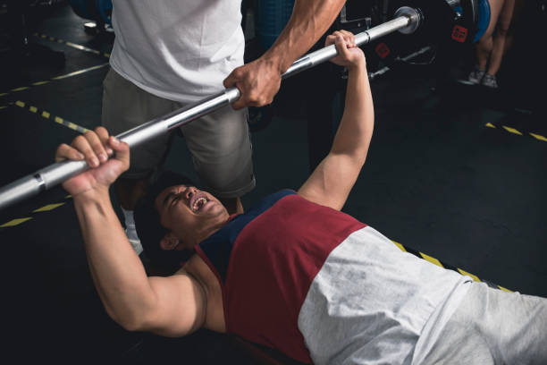 ein junger mann, der bankdrücken macht, schaltet sich durch eine letzte wiederholung ein, während ein anderer kerl ihn entdeckt. brusttraining im fitnessstudio. - spotting stock-fotos und bilder