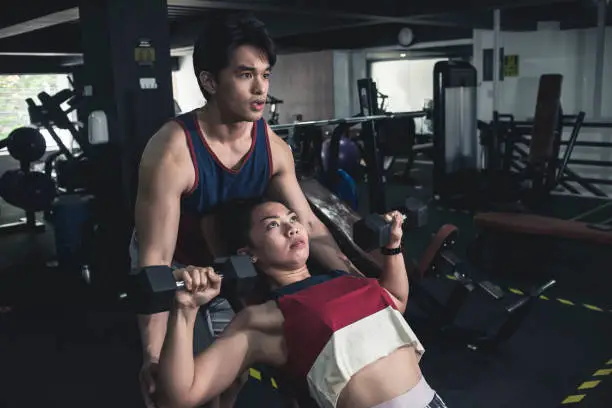 A young asian man spotting an athletic girl doing incline dumbbell presses. Working out with assistance at the gym.