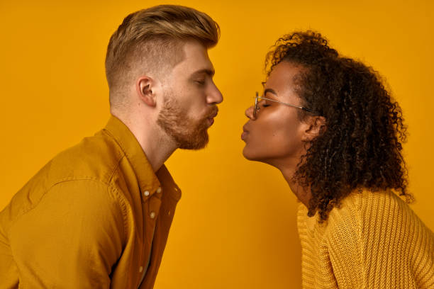 istock Mixed race couple stand in profile, keep lips folded, staring at each other, going to kiss, dressed casual mustard color wear, make grimace, pose over orange wall, have fun. Facial expressions 1367619840