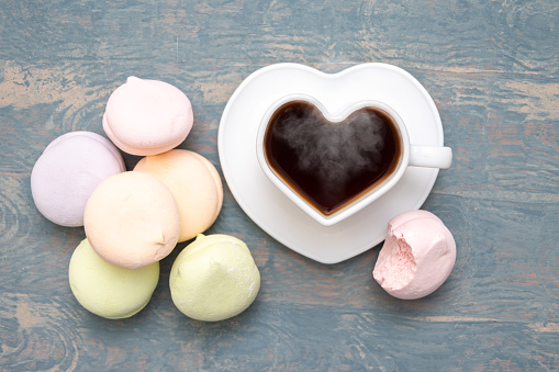 Round multi-colored marshmallows in a pile, a bitten off half of a marshmallow and a white heart-shaped mug with black steaming hot coffee on a white plate on a blue wooden background