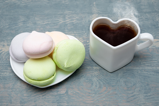 Round multi-colored marshmallows in a pile and a white heart-shaped mug with black steaming hot coffee on a blue wooden background. Breakfast for loved ones.